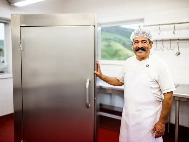 Smiling meat market owner in a white apron and hairnet resting a hand on a stainless-steel smokehouse cabinet
